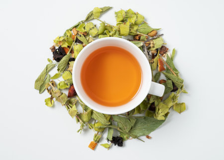 A cup of herbal tea with dried herbs scattered around and pieces of fruit on a white background. Top view and copy space.の写真素材