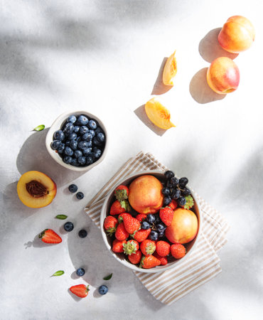 Fresh organic summer berries and fruits of strawberries, blueberries, peaches and grapes in a white plate on a light background with morning shadow. Top view and copy space.の写真素材