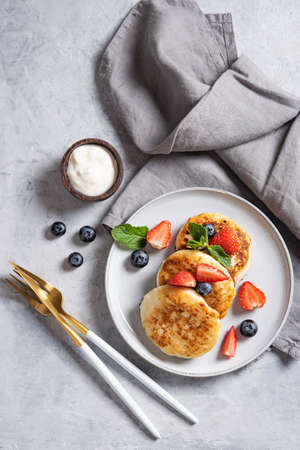 Traditional Russian dish for breakfast  syrniki. Cottage cheese pancakes with sour cream and fresh berries on a blue background with tableware and napkin. Top view image. Vertical orientation.の写真素材
