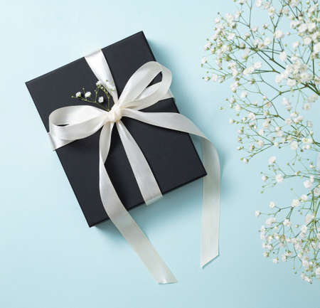 a black gift box with a white ribbon on a blue background with a bouquet of  gypsophila. The concept of a holiday or gift card. Top  view and copy space.の写真素材
