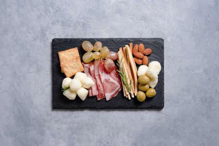 A plate with cheese, ham, mozzarella, olives, nuts, grapes and other snacks on a black stone board on blue background. The concept of Italian cuisine. Top view and copy space.の写真素材
