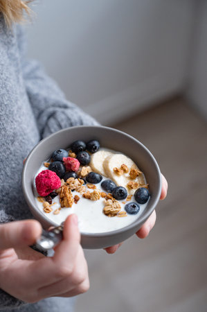 a young girl in a gray sweater is holding a bowl of vegetarian yogurt, granola and fresh fruit on...