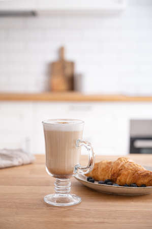 Glass of fragrant cappuccino coffee with cinnamon and croissant on a wooden  table against the background of a white kitchen in the early morning. Breakfast concept. Front   
view.の写真素材