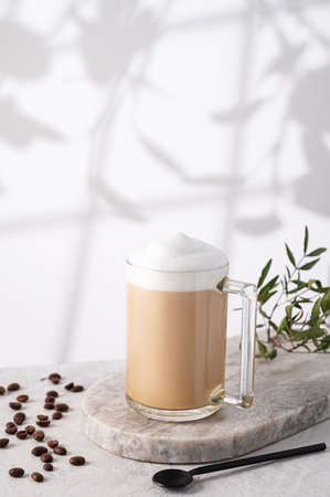 Cappuccino or latte with milk foam in a cup with coffee beans and spoon  on light  marble background with morning shadow.の写真素材