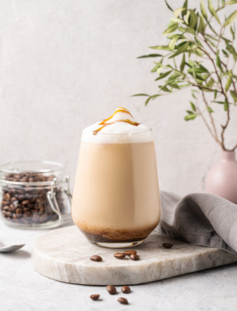 Cappuccino or latte with milk foam and caramel in a glass with coffee beans on light  marble background with branches. Front view and copy space.の写真素材