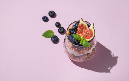 Delicious natural yogurt with granola, figs and blueberries in a glass on a pink background with...