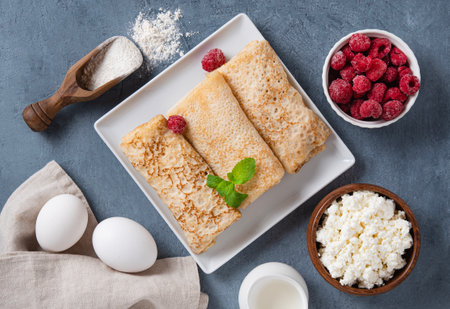Homemade crepes with raspberry and cottage cheese on a plate on a blue background with eggs and flour. Healthy food for the breakfast concept. Top view.の写真素材