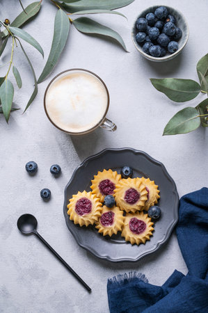 Cappuccino or latte coffee with milk foam in a cup with  homemade berry cookies in a dark plate and blueberries on a light background with eucalyptus branches. Concept spring morning breakfast. Top view.の写真素材