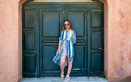 A girl in a beautiful summer dress and sunglasses is standing near an old wooden green door on a sunny day. Summer trip to ancient sights in Corfu. Greece.の写真素材