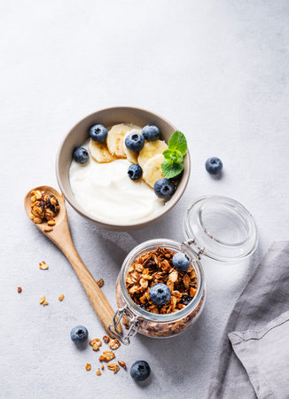 Homemade baked granola with yogurt, blueberries and banana in a bowl on a light background with fresh berries. Healthy vegetarian breakfast with muesli. Top view and copy space.の写真素材