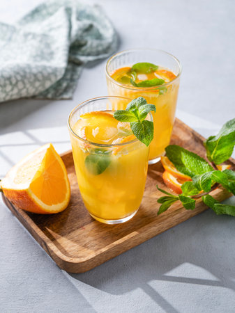 Freshly squeezed juice in a glass with fresh oranges, mint and ice on a light background with metallic tube and shadow. Healthy citrus detox drink for breakfast. Copy space.の写真素材
