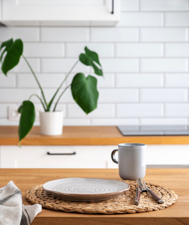 Wooden table top with plate and cup for breakfast against blurred white kitchen with cutting board and monstera plant in scandinavian style in morning lightの写真素材