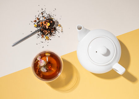Creative flat lay of a glass cup of herbal tea with rose flowers on a yellow and white background with teapot and hard shadow. Food and drink concept. Top view and copy space.の写真素材