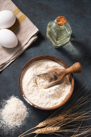 Wheat flour in a wooden bowl with a scoop on a dark blue background with eggs, oil and dry branches wheat. Organic ingredients for baking. Homemade healthy food concept. Top view.の写真素材