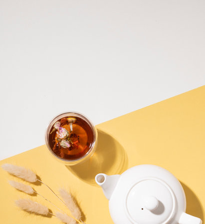 Creative layout of a glass cup of herbal tea with flowers and fruits and teapot on a white and yellow background with a shadow. Healthy drink concept. Top view and copy space.の写真素材