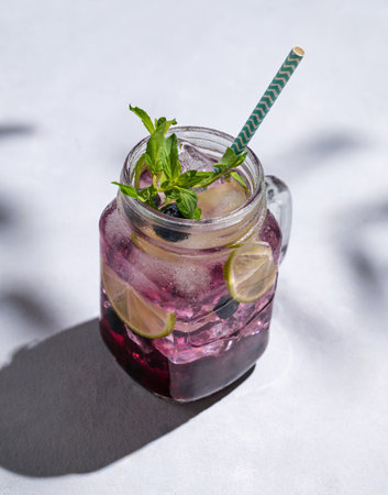 Blueberry mojito or lemonade with lime, ice and mint in a glass on a light background with berries, fruit and shadows. The concept of a summer refreshing drink. Free space for text.の写真素材