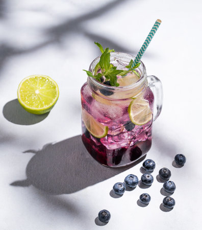 Blueberry mojito or lemonade with lime, ice and mint in a glass on a light background with berries, fruit and shadows. The concept of a summer refreshing drink. Top view and copy space.の写真素材