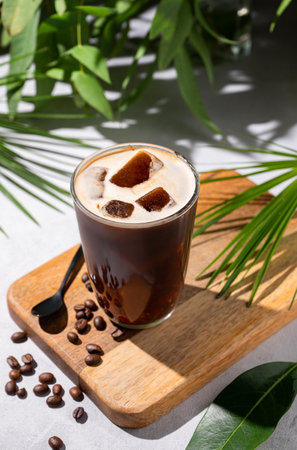 Iced coffee brew in a glass on a wooden board on a light background with coffee beans and morning shadows in tropical branches. Summer refreshment concept. Top view.の写真素材