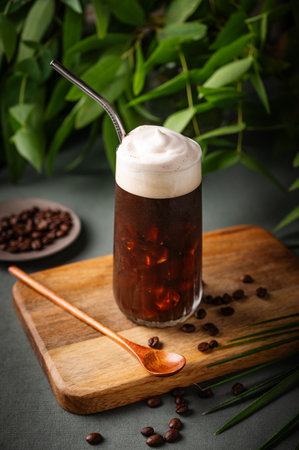 Iced coffee brew with foam  in a glass on a wooden board on a green background with coffee beans in tropical branches. Summer refreshment concept.の写真素材