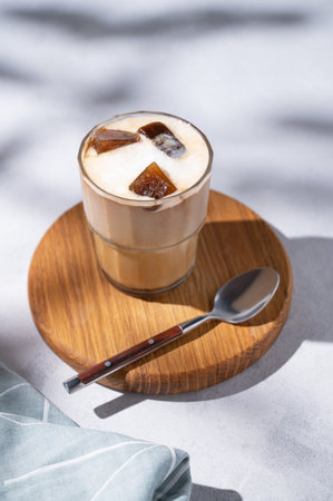 Iced coffee latte in a glass with milk on a wooden board on a light background with spoon, coffee beans and morning shadows. Summer refreshment concept. Free space for text.の写真素材