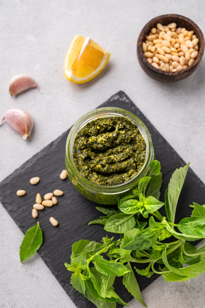 Traditional Italian pesto in a jar with green basil, pine nuts, cheese and olive oil on a slate board on a light background. A classic sauce for spaghetti or bruschetta. Top view.の写真素材