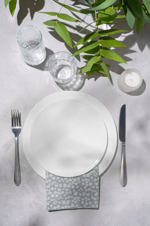 Festive table setting with white plates, glasses and candle for a celebration with eucalyptus branches on a light background with shadows. Restaurant menu concept. Top view.の写真素材