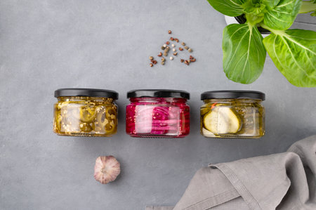 Pickled jalapeno peppers, red onions and cucumbers in jars on a blue background with fresh herbs and spices. Traditional canned homemade snacks. Healthy fermented food. Top view and copy space.の写真素材