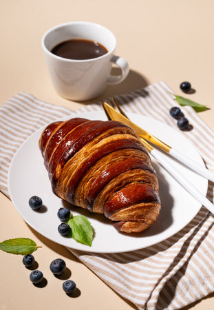 Freshly baked croissant with chocolate on a white plate and blueberries on a yellow background with cup of black coffee. The concept of a delicious homemade breakfast.の写真素材