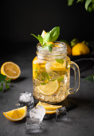 Lemonade drink with fresh lemon, mint and ice in a jar. Refreshing citrus mojito cocktail or cold tea on a dark background. Homemade healthy and detox drink concept. Front view.の写真素材