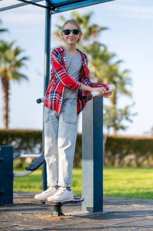 A joyful girl in a plaid shirt is training on exercise equipment in a park outdoors on a sunny summer day. The concept of sports and healthy lifestyle for a teenager.の写真素材