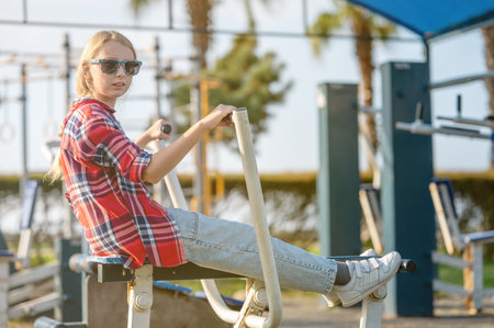 A joyful girl in a plaid shirt is training on exercise equipment in a park outdoors on a sunny summer day. The concept of sports and healthy lifestyle for a teenager.の写真素材