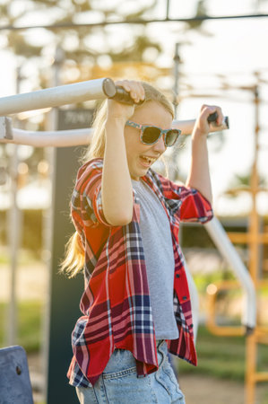 A joyful girl in a plaid shirt is training on exercise equipment in a park outdoors on a sunny summer day. The concept of sports and healthy lifestyle for a teenager.の写真素材
