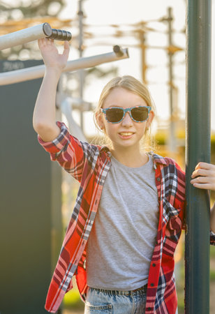A joyful girl in a plaid shirt is training on exercise equipment in a park outdoors on a sunny summer day. The concept of sports and healthy lifestyle for a teenager.の写真素材