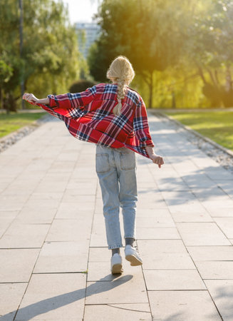 A girl in a plaid shirt jumps and runs along a path in a city park on a sunny summer day. Teenage holiday concept.の写真素材
