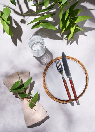Celebration table setting with a light empty plate, a glass, and a napkin with eucalyptus branches on a light background with shadows. Rustic style. Restaurant menu concept. Top view.の写真素材