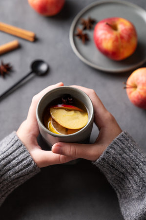 Hands hold a gray glass with homemade apple punch or cider with apples and cinnamon on a dark background with fresh fruits and spices. The concept of an autumn or winter healthy drink. Top view.の写真素材