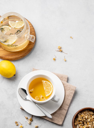 Chamomile herbal tea with lemon in a white cup and teapot with flowers on a light background. The...
