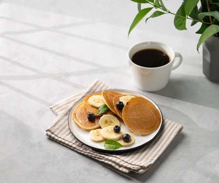 Banana pancakes with fresh fruit slices, berries, mint and honey on a light background with a cup of black coffee, shadows and green branch. Healthy diet breakfast. Free space for text.の写真素材