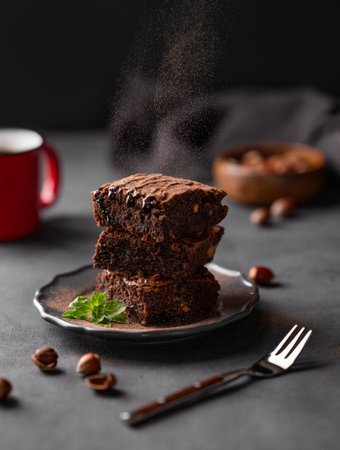Cocoa pours on to a stack of chocolate brownies with hazelnuts and salted caramel on a dark background with a cup of coffee and nuts. The concept of delicious and sweet homemade baked goods.の写真素材