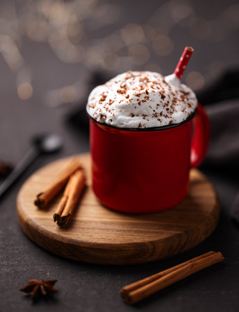 Red mug with hot chocolate or cocoa with whipped cream on a wooden stand with cinnamon sticks and star anise scattered around on a dark background with a garland in blur. Winter mood concept.の写真素材