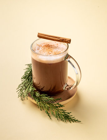 Glass mug of hot chocolate with whipped cream and cinnamon stick on a stand with pine branches on a yellow background. Concept for a winter warming Christmas drink. Free space for text.の写真素材