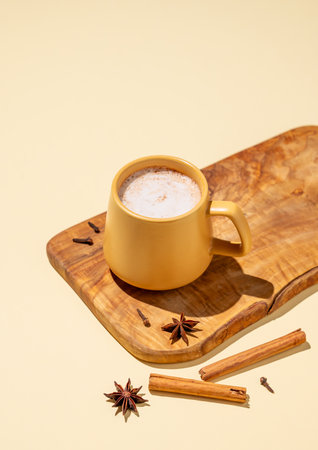 Masala latte tea. Traditional Indian drink with a mixture of spices and herbs. A yellow mug with a frothy drink surrounded by cinnamon sticks and star anise on a wooden board on a yellow background.の写真素材