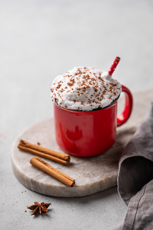Red mug of hot cocoa or chocolate with whipped cream and cinnamon sticks on a gray background. The mug sits on an oval marble stand with a napkin. Free space for text.の写真素材