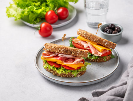 Two toast club sandwiches made of grain bread with cheddar cheese and bacon stuffed with tomatoes and lettuce and arugula on a light background with olives and glass of water.の写真素材