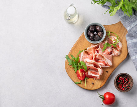 Flat lay of a wooden board with bacon or prosciutto on a light background with olives, spices, tomatoes and herbs. Antipasto concept. Top view and copy spaceの写真素材