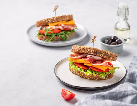 Two toast club sandwiches made of grain bread with cheddar cheese and bacon stuffed with tomatoes and lettuce and arugula on a light background with olives and oil. Healthy and dietary breakfastの写真素材
