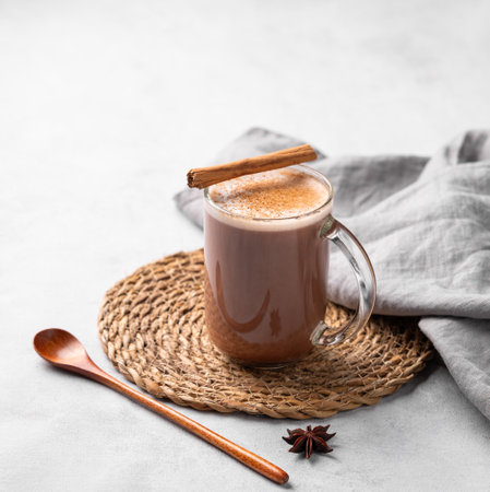 Glass mug with hot chocolate and milk foam on a light background with cinnamon sticks, anise star and spoon. Warming winter  drink concept. Free space for text.の写真素材