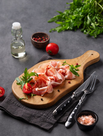 Snack wooden board with bacon or prosciutto on a dark background with spices, tomatoes and herbs. Antipasto concept.の写真素材