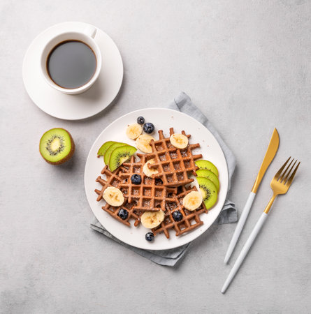 A freshly prepared chocolate waffles with kiwi, blueberries and banana slices on a white plate with a cup of coffee on a light gray background. The concept of a healthy dietary and delicious breakfastの写真素材