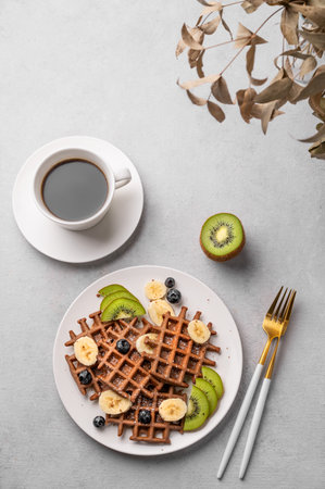 Vegan belgian waffles with kiwi, blueberries and banana slices on a white plate with a cup of coffee on a light background. The concept of a healthy dietary and delicious breakfast. Top view.の写真素材